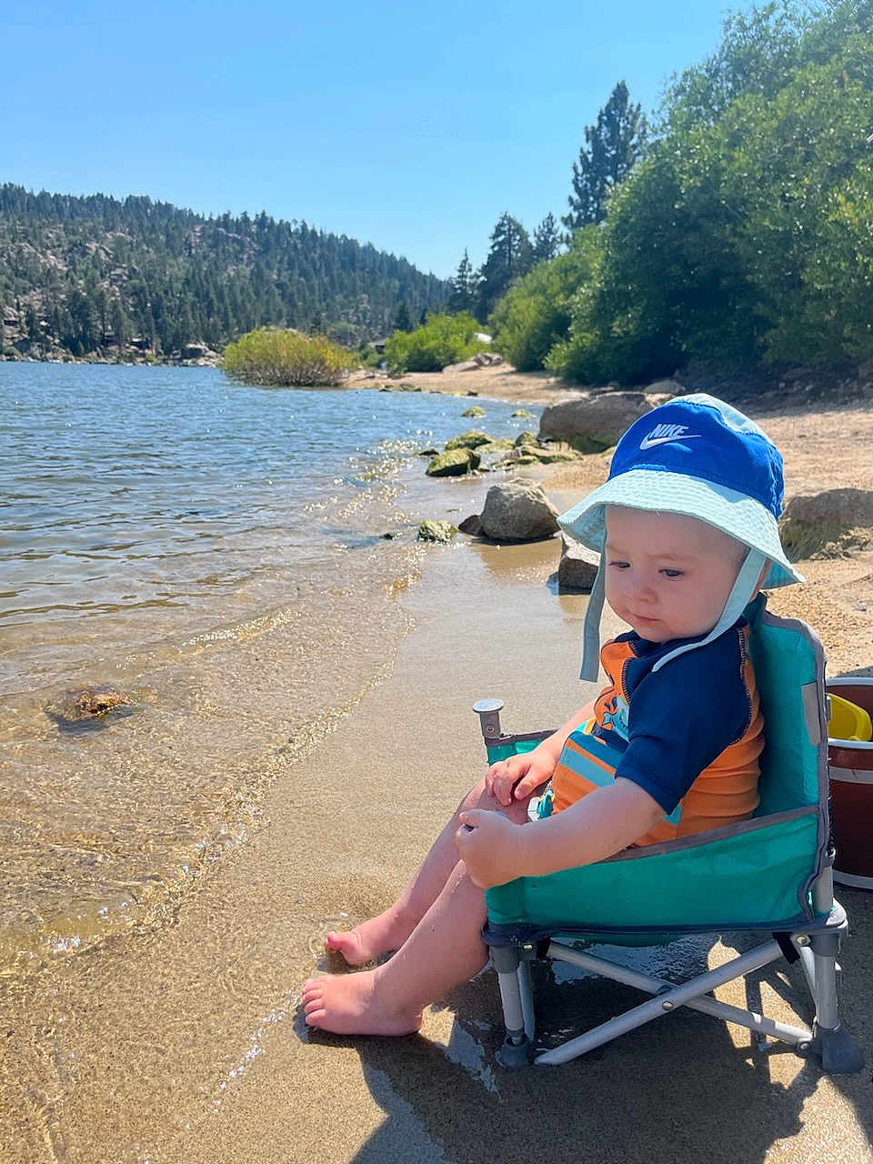 Caydence is registered to the contest to win money with this photo: toddler, child, beach_chair, water, lake, sand, hat, blue_sky, forest, trees, rocks, sunlight, outdoor, nature, summer, swimsuit, footwear, reflection, vacation, relaxation