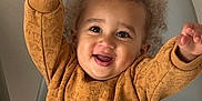 Kaylon a rejoint le concours — aidez-le/la à gagner de superbes lots ! brown_sweater, casual_clothing, chair, cheerful, child, curly_hair, cute, grey_pants, happy, home, indoor, person, playful, portrait, raised_hands, sitting, smiling, sweater, toddler, young_child