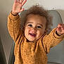 brown_sweater, casual_clothing, chair, cheerful, child, curly_hair, cute, grey_pants, happy, home, indoor, person, playful, portrait, raised_hands, sitting, smiling, sweater, toddler, young_child