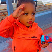 Ousman participe au concours pour gagner de l'argent avec cette photo : child, orange_hoodie, toy_truck, sunglasses, train_station, blue_train, platform, curious, standing, casual_clothing, outdoor, daylight, person, playing, transportation, waiting, young_child, holding_toy, urban, background