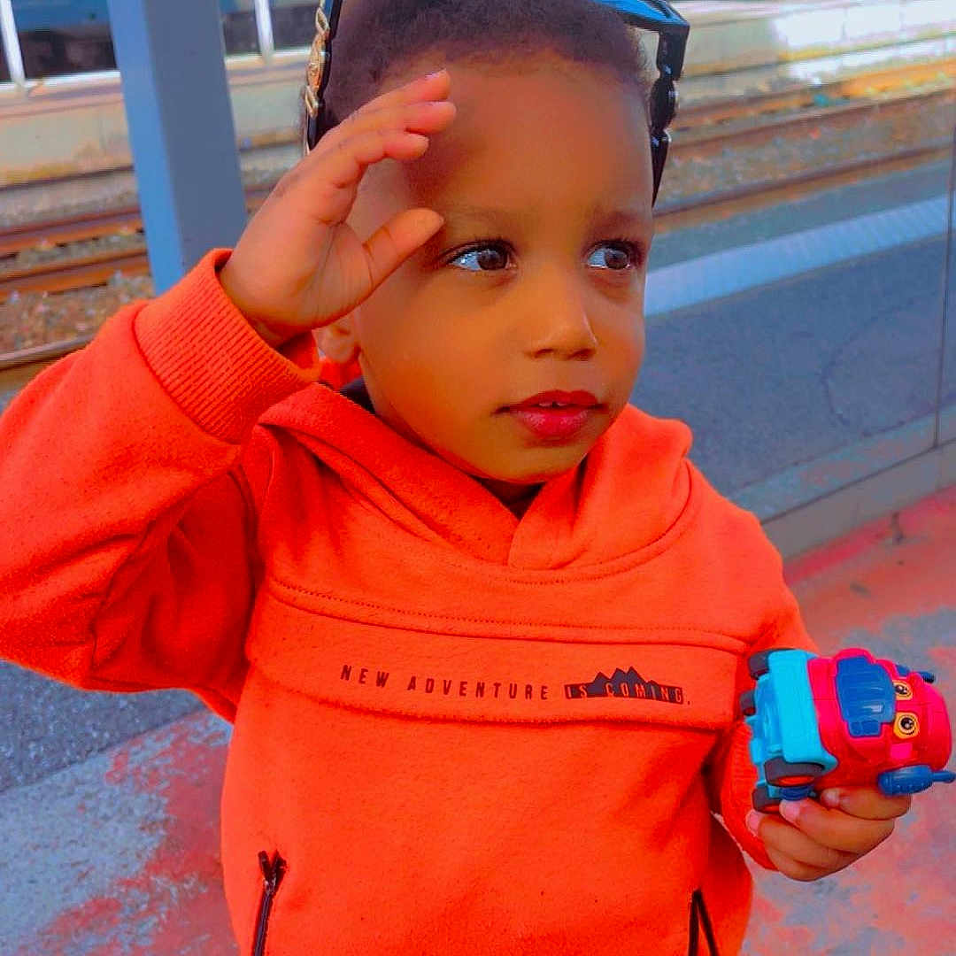 Ousman participe au concours pour gagner de l'argent avec cette photo : background, blue_train, casual_clothing, child, curious, daylight, holding_toy, orange_hoodie, outdoor, person, platform, playing, standing, sunglasses, toy_truck, train_station, transportation, urban, waiting, young_child