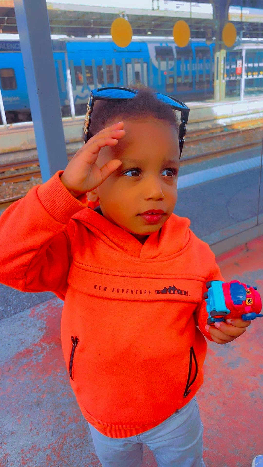 Ousman participe au concours pour gagner de l'argent avec cette photo : child, orange_hoodie, toy_truck, sunglasses, train_station, blue_train, platform, curious, standing, casual_clothing, outdoor, daylight, person, playing, transportation, waiting, young_child, holding_toy, urban, background
