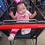 baby, shopping_cart, pink_bow, pink_clothing, socks, curly_hair, indoor, floor, clothing_racks, metal, shopping, child, person, retail, store, cart_handle, expression, footwear, seat, shopping_area