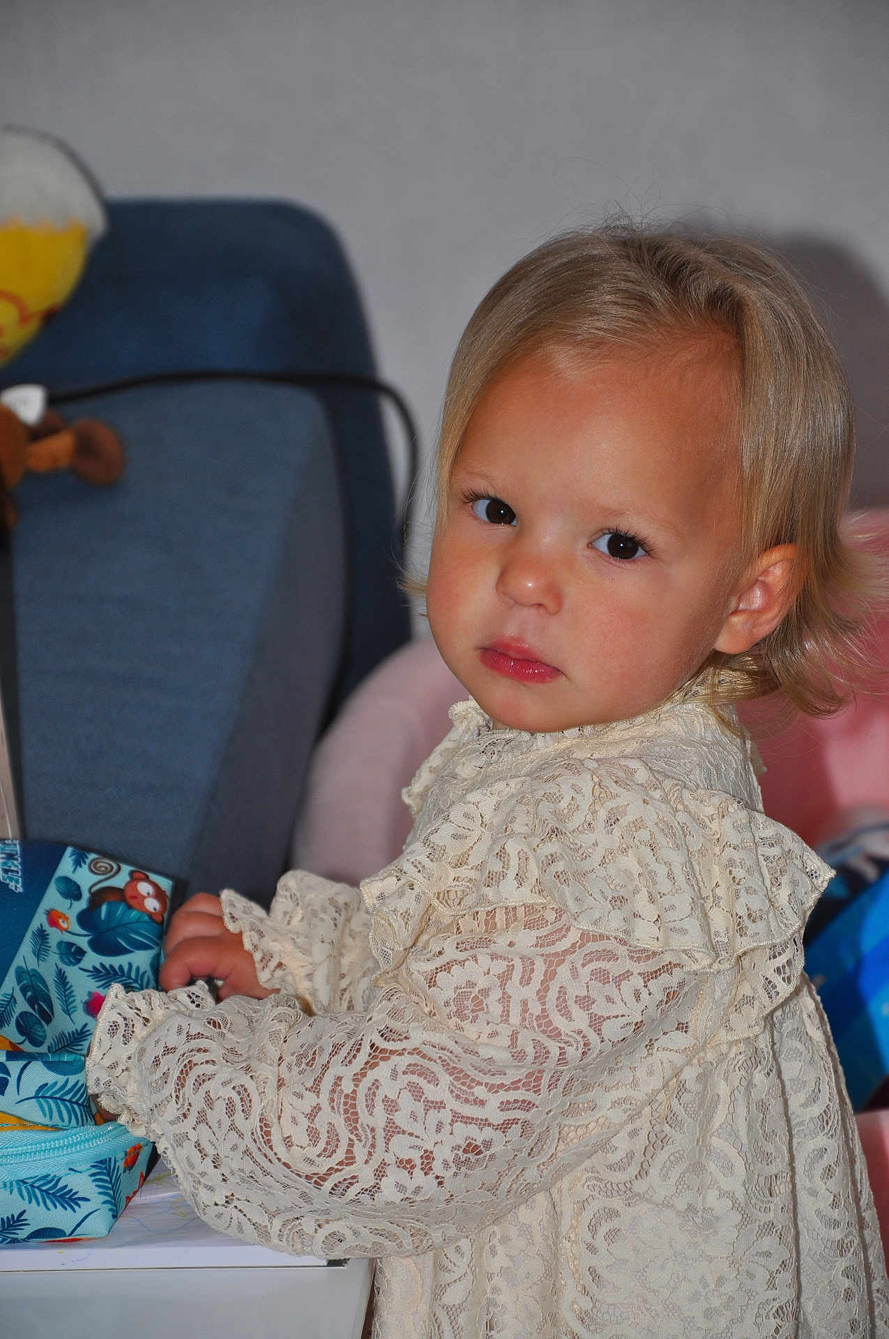 Ameliia participe au concours pour gagner de l'argent avec cette photo : toddler, child, blonde_hair, lace_clothing, indoor, serious_expression, furniture, blue_chair, colorful_bag, curious, portrait, young_child, person, cute, soft_lighting, home, table, sitting, face, closeup