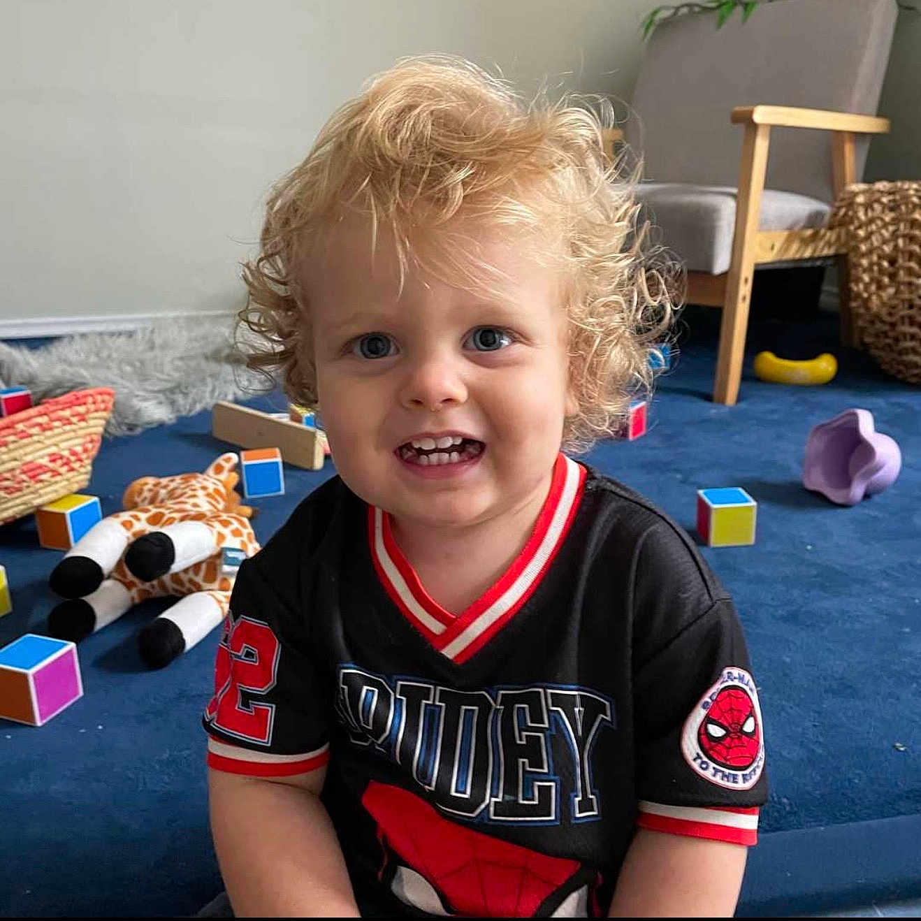Korrowa is registered to the contest to win money with this photo: armchair, black_shirt, blonde_hair, blue_carpet, blue_eyes, child, colorful_blocks, curly_hair, cute, happy, indoor, playroom, portrait, smile, spiderman_shirt, teeth, toddler, toy_giraffe, toys, woven_basket