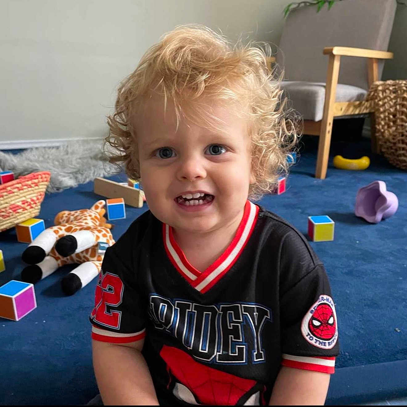 Korrowa is registered to the contest to win money with this photo: child, toddler, curly_hair, blonde_hair, blue_eyes, smile, teeth, spiderman_shirt, black_shirt, toy_giraffe, colorful_blocks, blue_carpet, playroom, armchair, woven_basket, indoor, portrait, happy, cute, toys