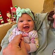 Everly is registered to the contest to win money with this photo: baby, infant, smiling, headband, hand_holding, couch, person, child, indoor, holiday_decor, santa_figure, happy, face, clothing, adult_hand, cute, portrait, cozy, home, family