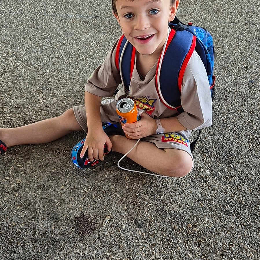 Luguy participe au concours pour gagner de l'argent avec cette photo : backpack, boy, casual_clothing, child, concrete_floor, cute, daylight, footwear, happy, orange_soda_can, outdoor, playful, portrait, short_sleeve_shirt, shorts, sitting, smiling, toy_story, watch, young