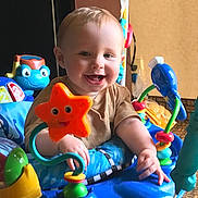 Daxton is registered to the contest to win money with this photo: baby, smiling, child, indoor, toy, activity_center, orange_star_toy, blue_fish_toy, colorful, happy, infant, play, plastic_toy, seat, person, cute, young_child, bright_colors, cheerful, early_childhood