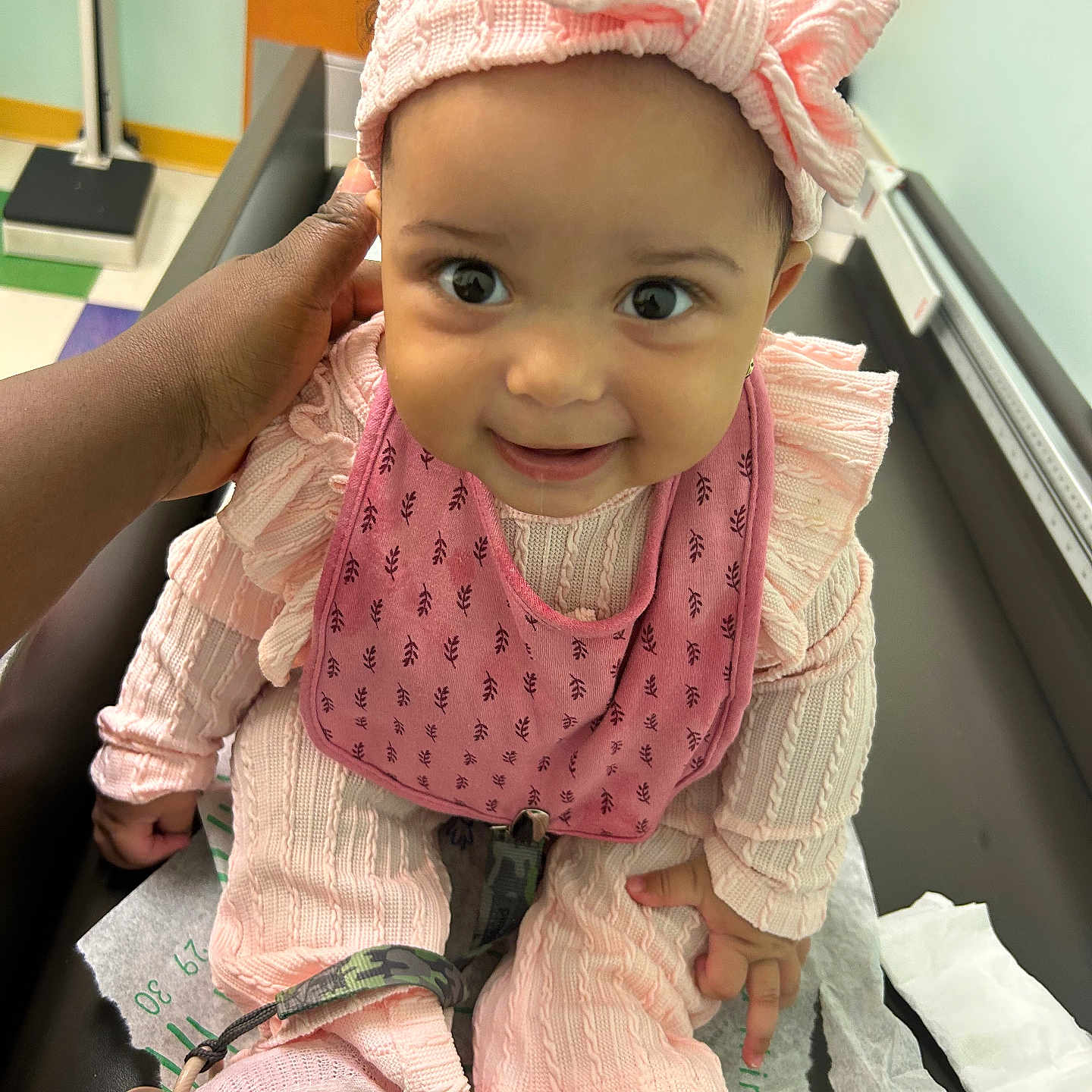 Magarteh is registered to the contest to win money with this photo: baby, bow, caregiver, child, closeup, cute, expression, floor_tiles, hand, headband, healthcare, hospital, indoors, infant, medical_table, pacifier_clip, person, pink_clothing, smile, socks
