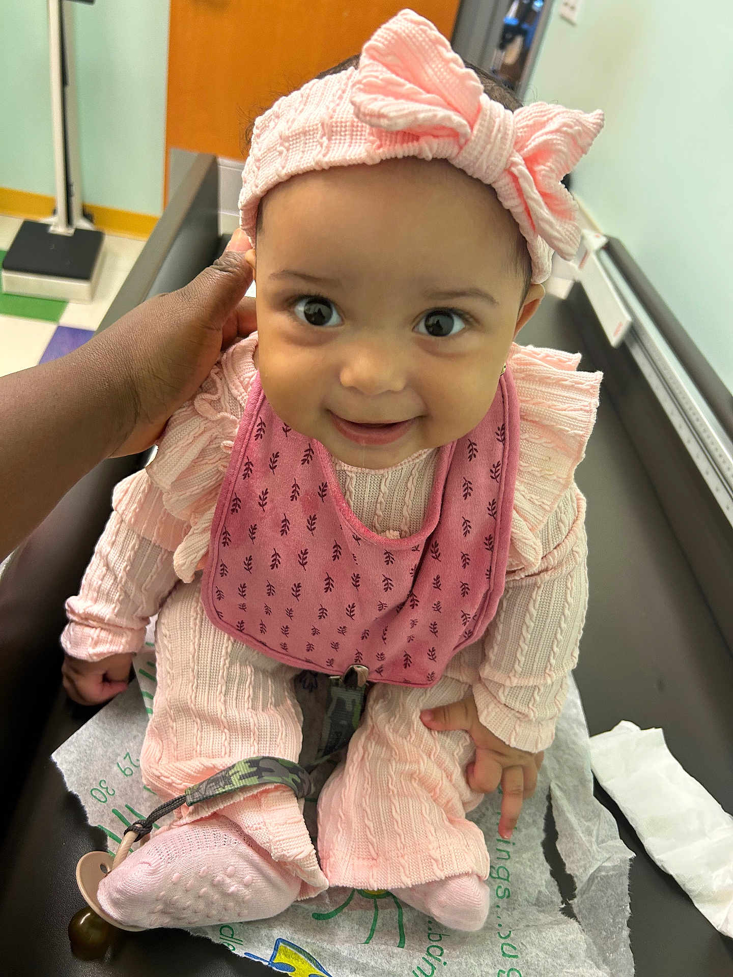 Magarteh is registered to the contest to win money with this photo: baby, infant, pink_clothing, headband, bow, smile, hand, medical_table, hospital, cute, child, socks, pacifier_clip, expression, person, indoors, floor_tiles, healthcare, caregiver, closeup