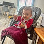 baby, high_chair, spoon, red_clothing, indoor, floor, table, curtains, window, computer, chair, desk, child, person, happy, feeding, snack, home, furniture, toddler