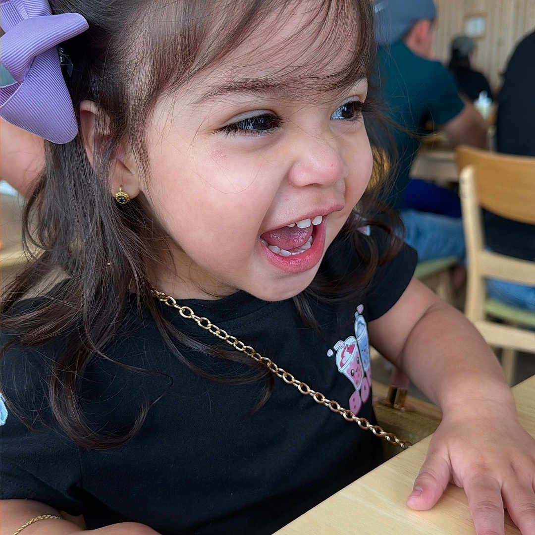 Maddie joined the competition — help win amazing prizes! child, girl, toddler, portrait, smiling, laughing, open_mouth, purple_bow, long_hair, earrings, black_shirt, necklace, hands, table, restaurant, cafe, indoor, wooden_chairs, warm_lighting, background_diners