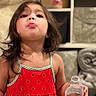 child, girl, toddler, portrait, indoors, red_dress, watermelon_pattern, water_bottle, kissy_face, long_hair, brown_hair, standing, shelves, stuffed_animal, couch, home, smiling, playful, close_up, photography