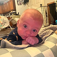 Caden is registered to the contest to win money with this photo: baby, infant, child, portrait, face, blue_eyes, looking_at_camera, resting, hands, blanket, checkered_blanket, bed, home, indoor, kitchen, baby_gear, warm_lighting, cozy, sitting, close_up