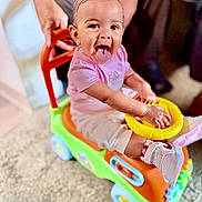 Dioř Aahmoř joined the competition — help win amazing prizes! baby, toddler, child, toy_car, pink_bow, playful, indoor, carpet, hand, person, sneakers, colorful, smiling, happy, cute, clothing, footwear, wheel, steering_wheel, parent