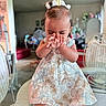 baby, child, curly_hair, cute, dress, face, floral, furniture, glass_table, hair_bows, hands, indoor, living_room, person, seated, shoes, smiling, socks, toddler, wicker_chair