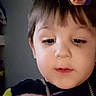 toddler, child, face, portrait, indoor, sweater, dinosaur, necklace, video_call, screen, adult, curly_hair, expression, casual_clothing, young_child, technology, communication, person, closeup, home