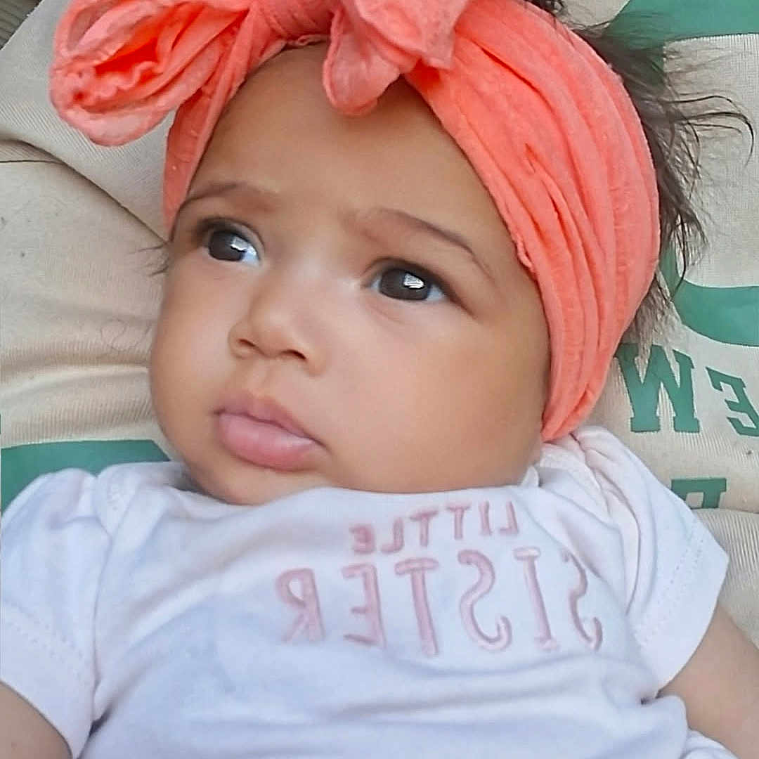 Heavenly joined the competition — help win amazing prizes! baby, child, infant, portrait, face, serious_expression, side_eye, headband, coral, bow, white_shirt, clothing, indoor, close_up, sitting, adult, caregiver, dark_hair, text_on_shirt, photography