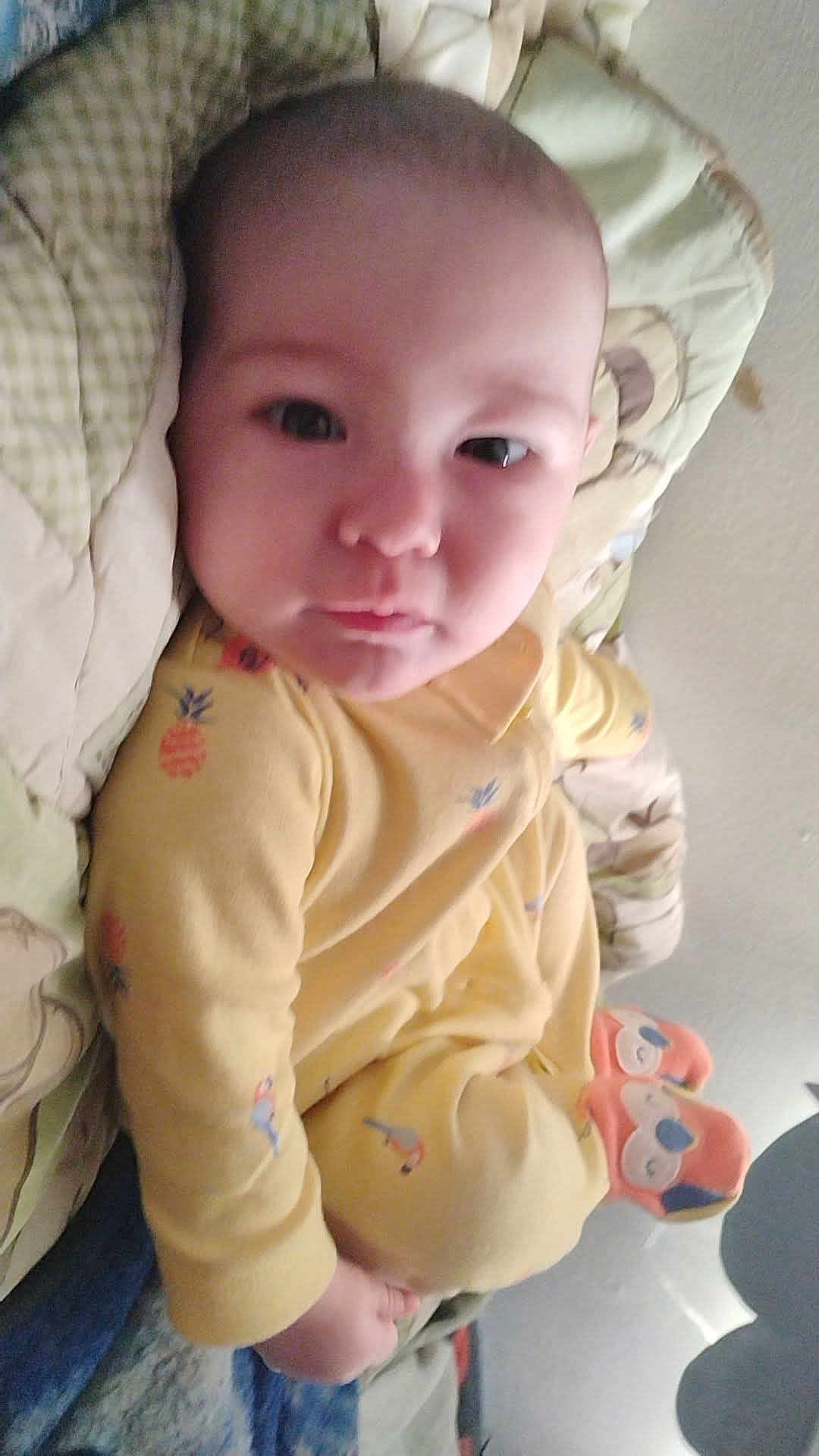 Ma'Kia Slapnicka is registered to the contest to win money with this photo: baby, child, infant, yellow_onesie, cushion, blanket, face, person, indoor, cute, expression, lying_down, soft_fabric, plush_toy, footwear, sleepwear, patterned_clothing, skin, head, eyes