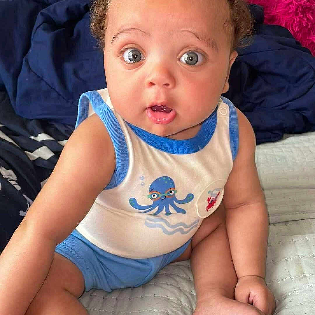 Tru is registered to the contest to win money with this photo: baby, bed, blanket, blue_shorts, child, curly_hair, cushion, cute, foot, hand, indoors, infant, octopus_print, person, sitting, skin, soft_fabric, surprised_expression, tank_top, wide_eyes