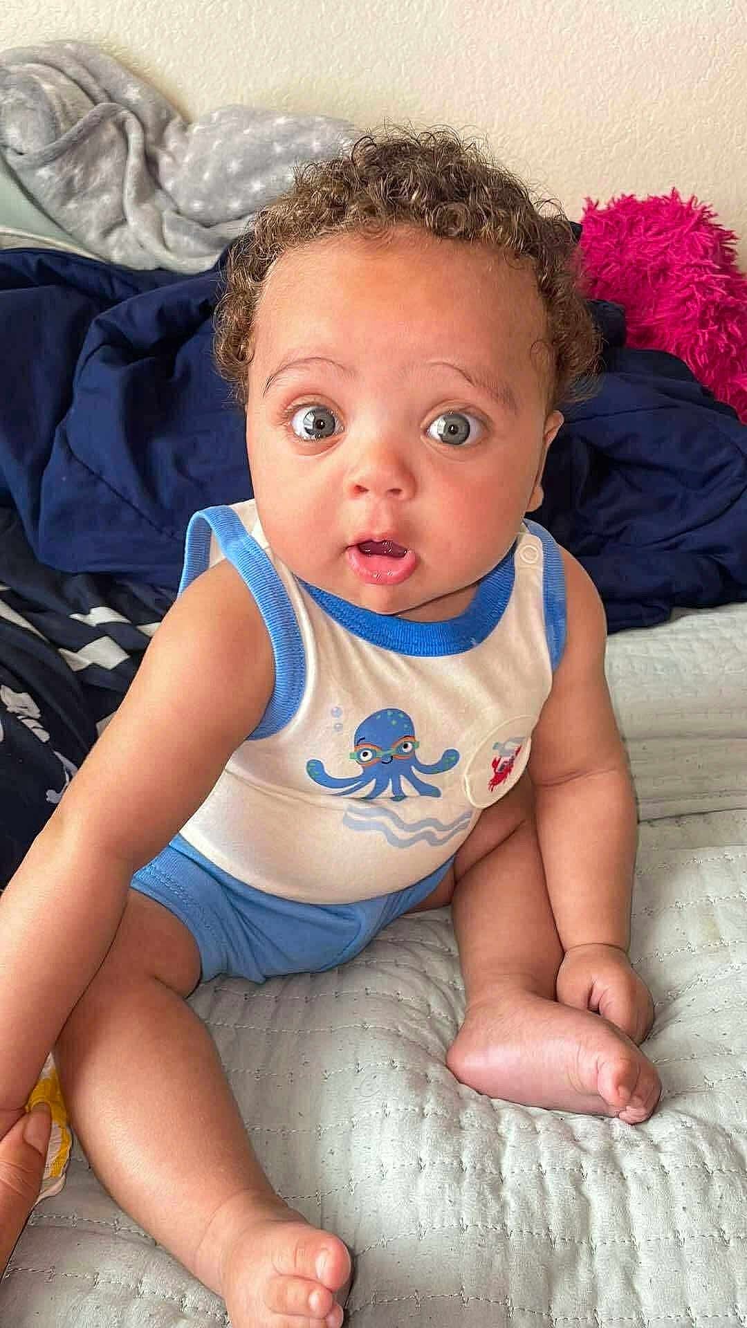 Tru is registered to the contest to win money with this photo: baby, child, curly_hair, blue_shorts, tank_top, octopus_print, bed, blanket, cushion, indoors, cute, wide_eyes, surprised_expression, skin, foot, hand, person, sitting, infant, soft_fabric