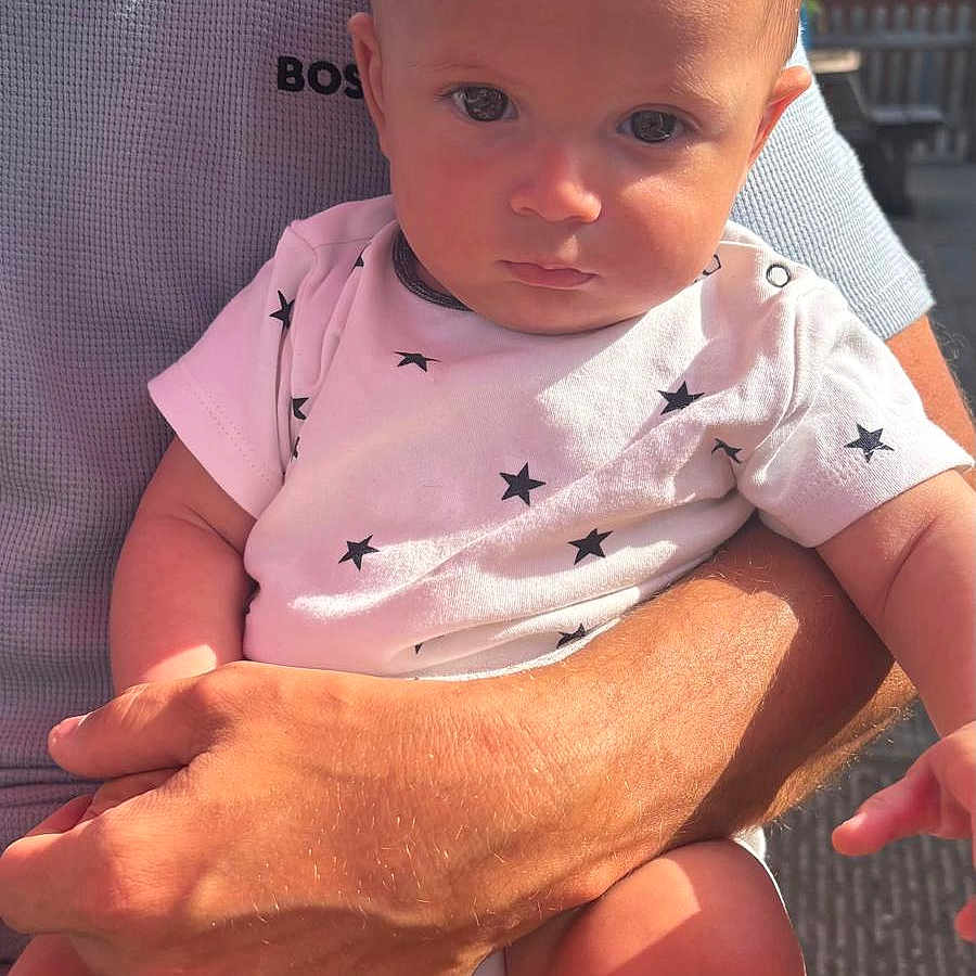 Archi-Lee is registered to the contest to win money with this photo: adult, arm, baby, child, closeup, clothing, comfort, expression, face, hand, holding, infant, onesie, outdoor, person, portrait, serious, skin, star_pattern, sunlight