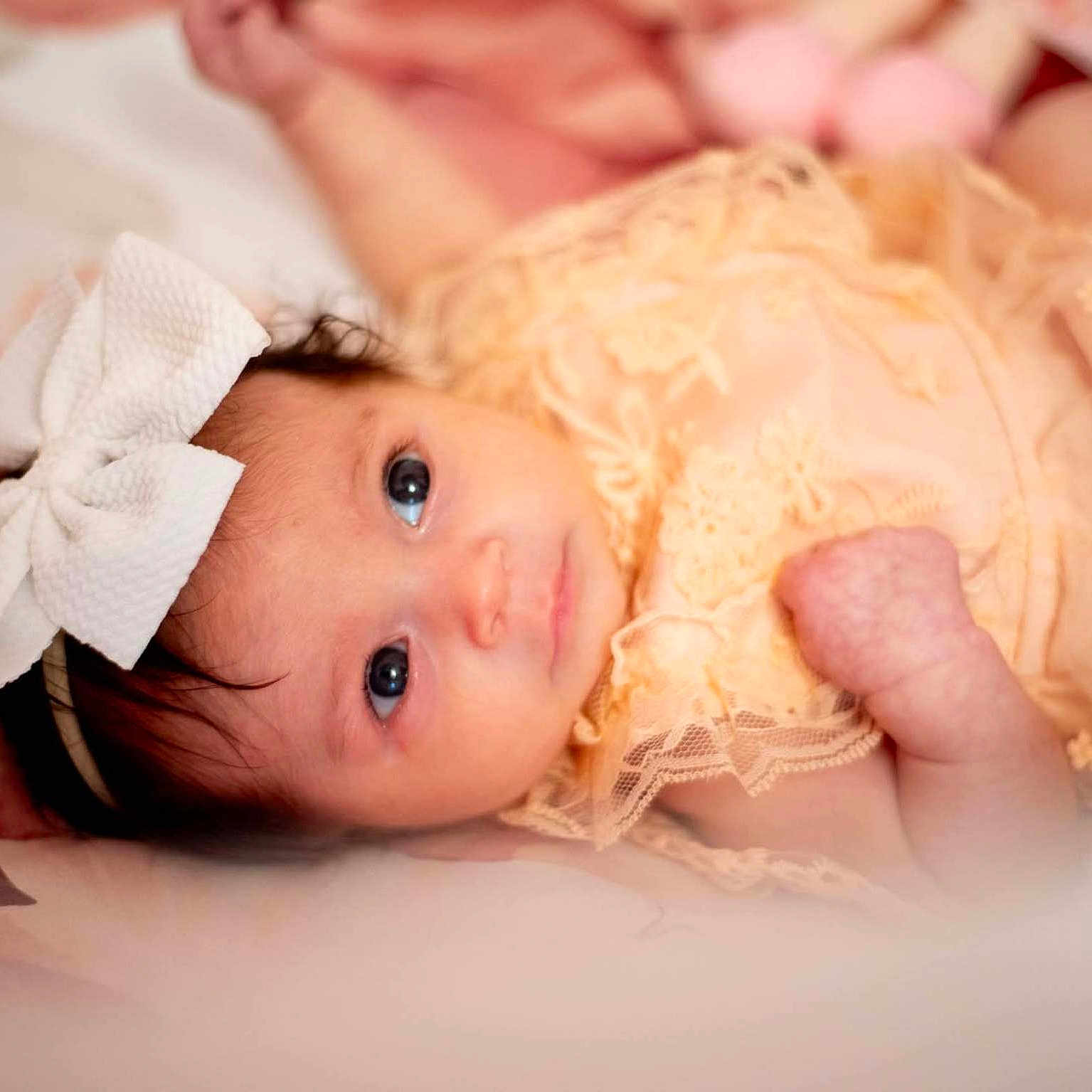 Scarlett is registered to the contest to win money with this photo: baby, infant, bow, headband, lace, peach, clothing, face, eyes, portrait, cute, newborn, soft_focus, lying_down, baby_girl, skin, expression, closeup, adorable, background