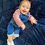 baby, child, smiling, happy, denim_overalls, striped_shirt, blue_eyes, pink_hair_ties, lying_down, soft_blanket, indoor, cute, infant, portrait, joyful, casual_clothing, white_socks, playful, baby_hair, cozy