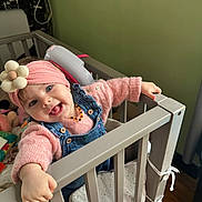 Eliana a rejoint le concours — aidez-le/la à gagner de superbes lots ! baby, child, crib, headband, pink_sweater, denim_overalls, flower_accessory, smiling, tongue_out, indoor, wooden_floor, baby_toys, happy, cute, portrait, person, playful, furniture, warm_clothing, home