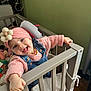 baby, child, crib, headband, pink_sweater, denim_overalls, flower_accessory, smiling, tongue_out, indoor, wooden_floor, baby_toys, happy, cute, portrait, person, playful, furniture, warm_clothing, home