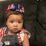 Jazaria is registered to the contest to win money with this photo: toddler, child, face, denim_overalls, bow, headband, red_white_blue, striped_shirt, sitting, indoor, couch, portrait, curly_hair, expression, cute, person, clothing, young_child, home, baby