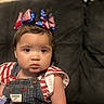 toddler, child, face, denim_overalls, bow, headband, red_white_blue, striped_shirt, sitting, indoor, couch, portrait, curly_hair, expression, cute, person, clothing, young_child, home, baby