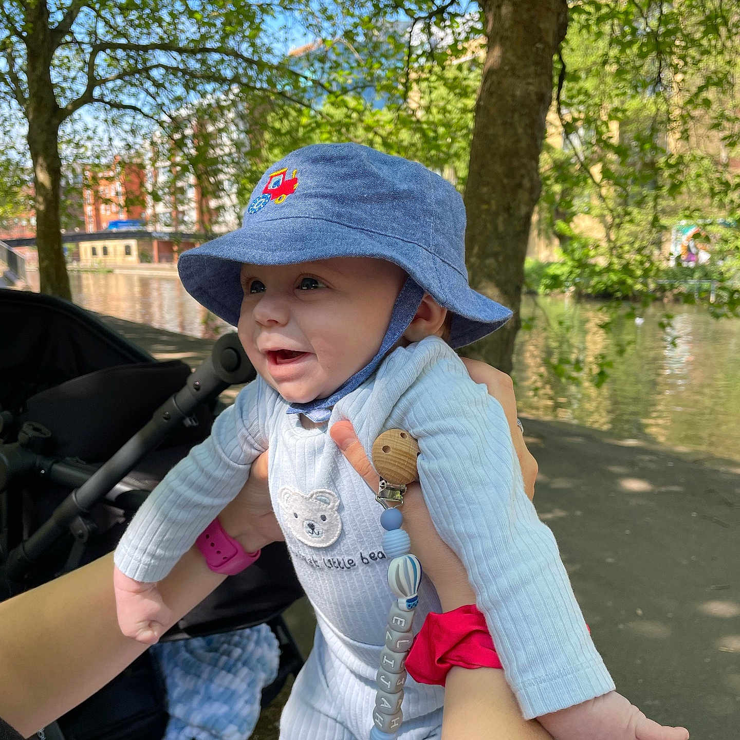 Elijah is registered to the contest to win money with this photo: baby, blue_hat, canal, child, cute, daylight, greenery, happy, holding_hands, nature, outdoor, pacifier_clip, park, person, playful, smiling, sunlight, trees, water, white_outfit