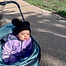 baby, child, stroller, outdoor, sidewalk, neighborhood, winter_clothing, hat, pom_poms, purple_jacket, blanket, infant, portrait, face, serious_expression, sunlight, park, pavement, walking, transportation