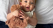 Sally is registered to the contest to win money with this photo: baby, child, infant, toddler, portrait, smiling, happy, bed, bedding, pillow, teddy_bear, stuffed_animal, headband, bow, white_outfit, indoor, bedroom, lying_down, hands, cuteness