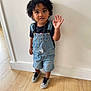 toddler, child, curly_hair, denim_overalls, sneakers, black_tshirt, wooden_floor, white_wall, standing, waving, cute, indoors, portrait, young_child, casual_clothing, person, flooring, expression, curly_hair_child, small_child
