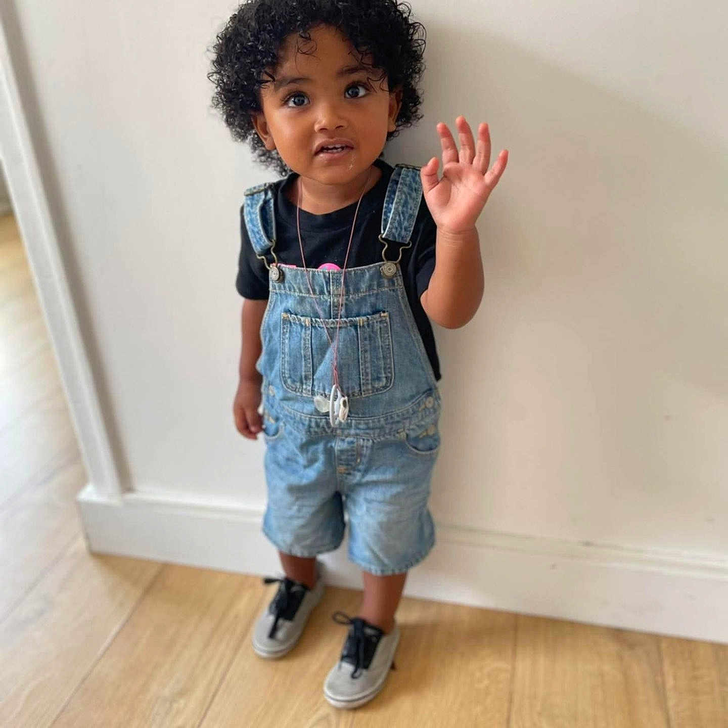 Yoënn a rejoint le concours — aidez-le/la à gagner de superbes lots ! black_tshirt, casual_clothing, child, curly_hair, curly_hair_child, cute, denim_overalls, expression, flooring, indoors, person, portrait, small_child, sneakers, standing, toddler, waving, white_wall, wooden_floor, young_child