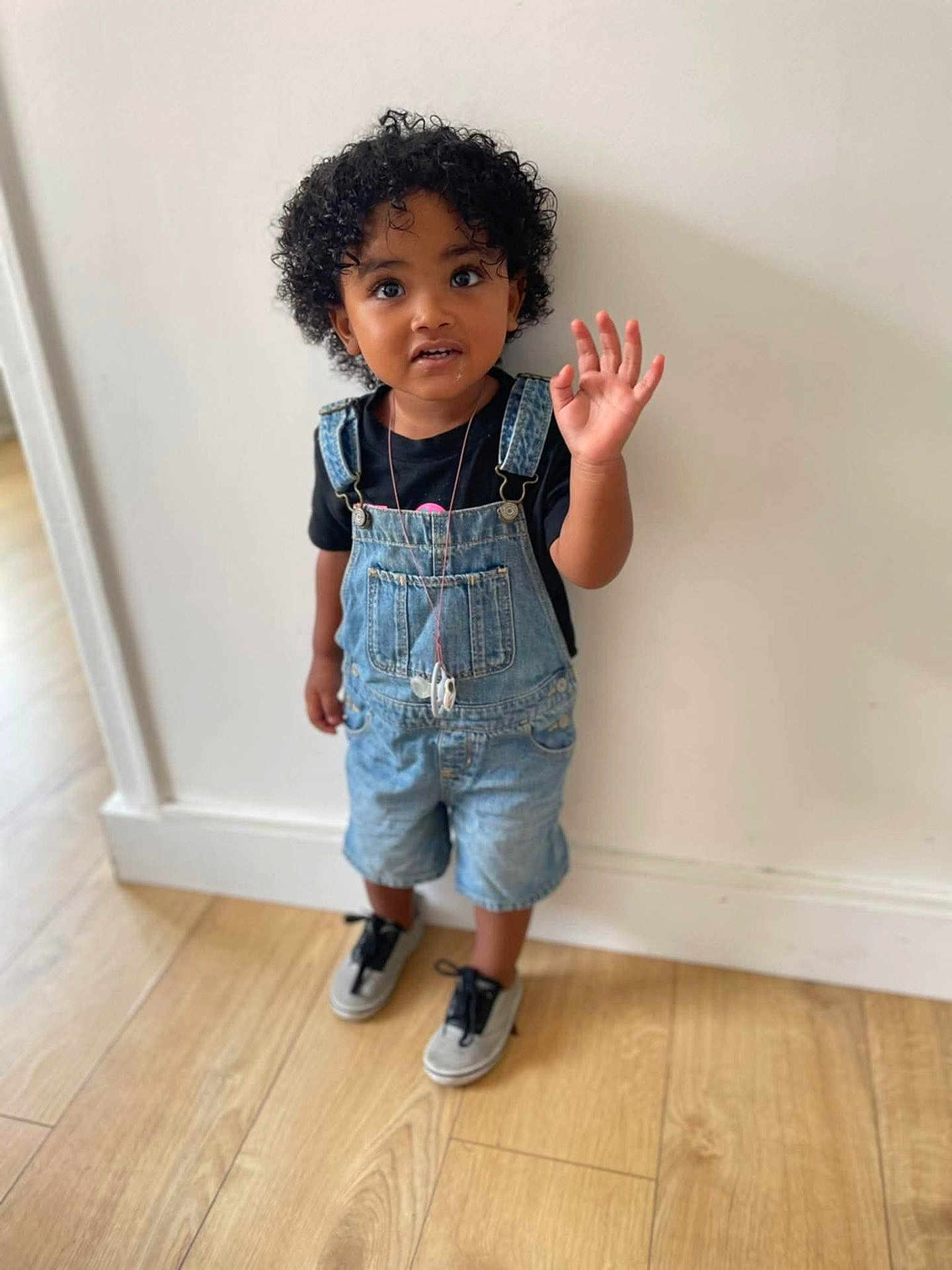 Yoënn a rejoint le concours — aidez-le/la à gagner de superbes lots ! toddler, child, curly_hair, denim_overalls, sneakers, black_tshirt, wooden_floor, white_wall, standing, waving, cute, indoors, portrait, young_child, casual_clothing, person, flooring, expression, curly_hair_child, small_child