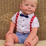 Finley is registered to the contest to win money with this photo: baby, child, sitting, barefoot, bow_tie, suspenders, striped_shorts, white_shirt, wicker_background, cushion, cute, portrait, blue_eyes, blond_hair, indoor, plump, smiling, person, fashion, toddler