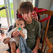 Armani is registered to the contest to win money with this photo: teenager, baby, pacifier, blanket, wooden_chair, curly_hair, indoor, window, glass_door, green_lawn, stroller, family, child, casual_clothing, floor_rug, holding, portrait, smile, person, home