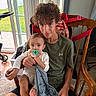 teenager, baby, pacifier, blanket, wooden_chair, curly_hair, indoor, window, glass_door, green_lawn, stroller, family, child, casual_clothing, floor_rug, holding, portrait, smile, person, home