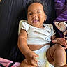 baby, infant, child, smiling, happy, portrait, lying_down, bed, blanket, purple, black, floral_clothing, diaper, short_sleeves, curly_hair, outdoorless, closeup, human, family, photography