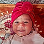 baby, smiling, red_hat, turban, face, child, infant, blanket, pattern, fabric, cute, portrait, happy, clothing, cozy, headwear, indoors, soft_texture, person, lying_down