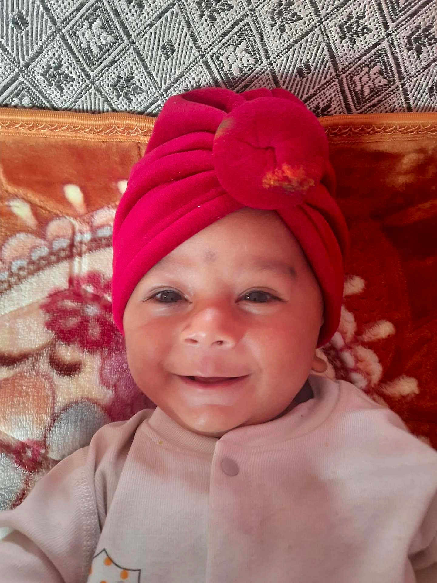 Saatvik joined the competition — help win amazing prizes! baby, smiling, red_hat, turban, face, child, infant, blanket, pattern, fabric, cute, portrait, happy, clothing, cozy, headwear, indoors, soft_texture, person, lying_down