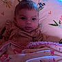 baby, blanket, cushion, floral_pattern, heart_pattern, wooden_wall, portrait, face, child, indoor, curly_hair, photography, soft_lighting, cozy, cute, expression, person, fabric, bedroom, decor