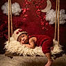A'Mere is registered to the contest to win money with this photo: baby, child, clouds, cozy, cute, decor, floor_rug, flowers, fluffy, hanging_flowers, indoors, infant, knitted_hat, portrait, red_outfit, resting, soft_texture, swing, warm_lighting, wooden_swing