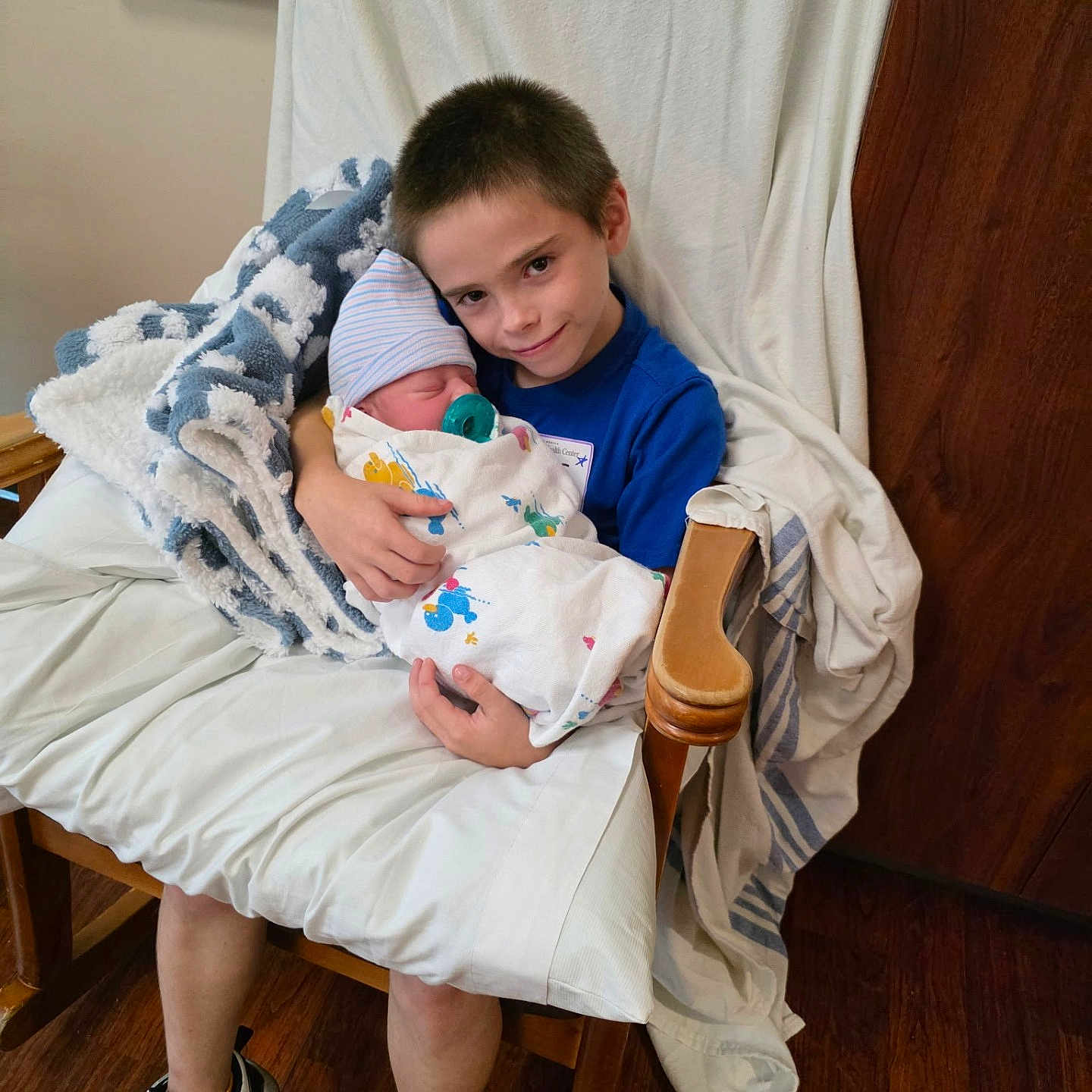 Alek is registered to the contest to win money with this photo: child, baby, newborn, boy, sibling, blanket, chair, cushion, indoor, wooden_floor, blue_shirt, pacifier, hat, holding, smile, family, tender_moment, seated, footwear, cozy