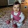 baby, child, smiling, sitting, pink_overalls, white_shirt, patterned_blanket, cookies, indoor, wooden_floor, kitchen, toys, hair, face, happy, clothing, person, floor, home, cute