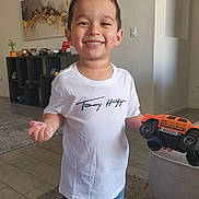 Axle is registered to the contest to win money with this photo: boy, cactus, carpet, casual_clothing, child, decor, floor_tiles, furniture, happy, home, indoor, living_room, monster_truck, person, portrait, shelf, shorts, smiling, toy, tshirt
